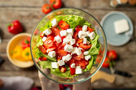 girls hands holding a vegetarian salad in a glass plate, sliced healthy food ingredients on a wooden table background, close-upの写真素材