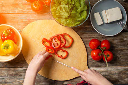 hands of a girl cut bell pepper on a wooden table, the process of making vegetarian salad, close-up cutting of vegetables and greensの写真素材
