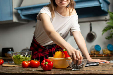 young cheerful girl prepares a vegetarian salad in the kitchen, she adds sliced ingredients to the...