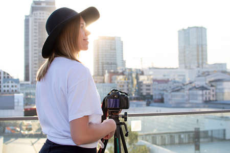 young girl photographer with a camera and a tripod on a background of the city, she photographs at sunset, a woman shoots a videoの写真素材