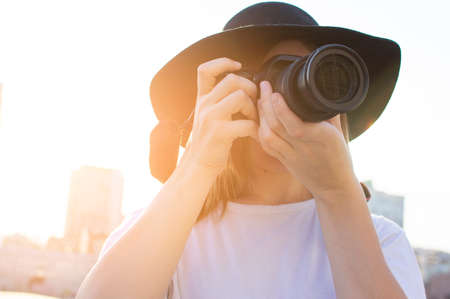 portrait of a young girl photographer, a woman with a modern camera photographs against the backdrop of a city sunsetの写真素材