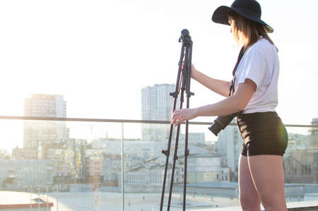 young girl-photographer with a camera and a tripod against the background of the city, she photographs the sunset, a woman getting ready for shooting, sets up a tripodの写真素材