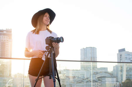 young girl photographer with a camera and a tripod on a background of the city, she photographs at sunset, a woman shoots a videoの写真素材