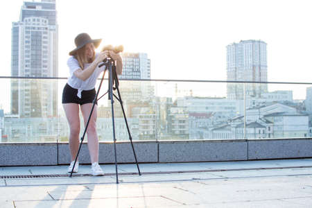 young girl photographer with a camera and a tripod on a background of the city, she photographs at sunset, a woman shoots a videoの写真素材