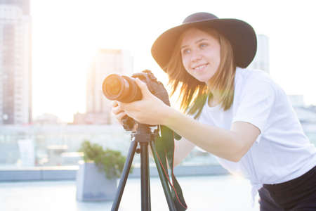 young girl photographer with a camera and a tripod on a background of the city, she photographs at sunset, a woman shoots a videoの写真素材