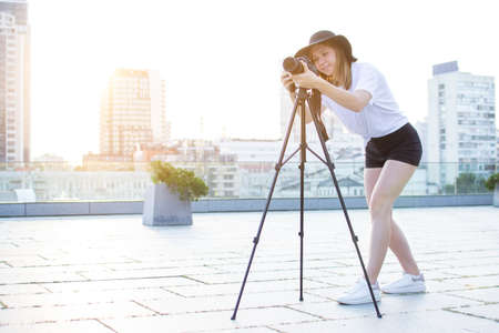 young girl photographer with a camera and a tripod on a background of the city, she photographs at sunset, a woman shoots a videoの写真素材