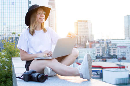 young girl photographer sitting with camera and laptop outdoors and retouching photo, tourist freelancer on a background of city sunsetの写真素材