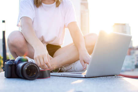 young girl photographer sitting with camera and laptop outdoors and retouching photo, tourist freelancer on a background of city sunsetの写真素材