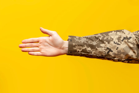 hand of a male soldier of the Ukrainian army in a camouflage pixel uniform shows a handshake and deal gesture on a yellow isolated background, empty palm of a military mans hand, close-upの写真素材