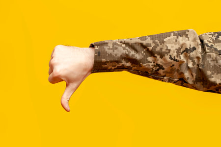 hand of a male soldier of the Ukrainian army in camouflage pixel uniform shows dislike and thumbs down on a yellow isolated background, bad gesture of failure and refusal, close-upの写真素材
