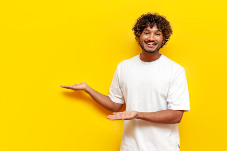 young curly indian man in a white t-shirt shows and advertises copy space on a yellow isolated background, a cheerful guy points with his hands to an empty spaceの写真素材
