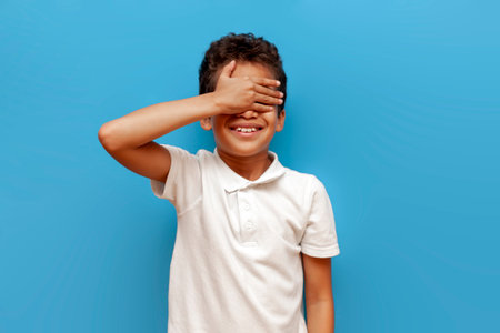 african american boy in a white polo closes his eyes with his hand and smiles on a blue isolated background, a 10 year old child waiting for a surpriseの写真素材