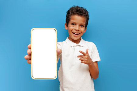 African-American boy in white polo shows blank smartphone screen on blue isolated background, 10-year-old child holds mobile phone and advertises copy spaceの写真素材