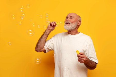 cheerful old bald grandfather in white t-shirt blowing soap bubbles on yellow isolated background, elderly pensioner playing with childrens toyの写真素材