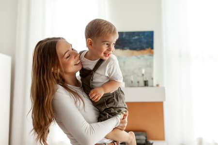 young mother holds her little son in her arms and lifts him up at home, 2-year-old boy is having fun...