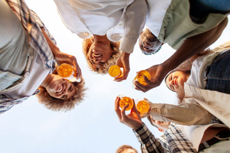 group of multiracial young people clink beer bottles outdoors, interracial students celebrating toast and relaxing together, six bottles of alcohol holding hands, bottom viewの写真素材