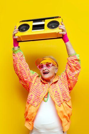old crazy grandmother in fashionable sportswear listens loudly to music on a tape recorder and dances on a yellow isolated background, an elderly funny pensioner with glasses raises up a boombox at a partyの写真素材