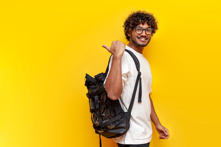 young indian guy student in a white t-shirt with glasses and with backpack points back on yellow isolated background, curly man goes to college to study and points at the copy space from behindの写真素材