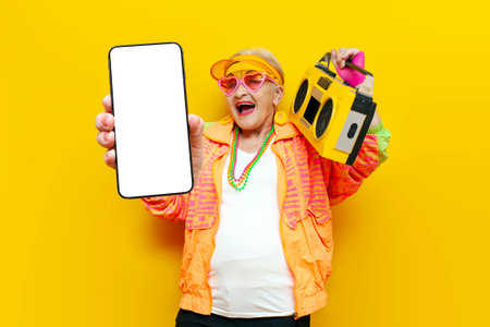 crazy cheerful grandmother with a tape recorder shows a blank screen of a smartphone on a yellow isolated background, an elderly pensioner woman listens to loud music online and uses the phoneの写真素材