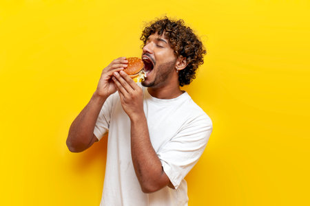 young indian man eating a big tasty burger with his mouth wide open on a yellow isolated background, curly guy student eating and advertising fast foodの写真素材
