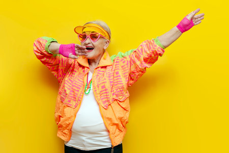 crazy funny old granny in sports colorful clothes and pink glasses dancing on a yellow isolated background, elderly woman in youth hipster clothes moving with raised handsの写真素材