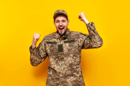 Ukrainian army soldier in pixel camouflage uniform celebrates victory and success on a yellow isolated background, Ukrainian military cadet winner rejoices in good luckの写真素材