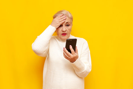 Confused old grandmother in a white sweater uses a smartphone and shows a facepalm on a yellow isolated background, sad elderly woman pensioner plays online on her mobile and worriesの写真素材