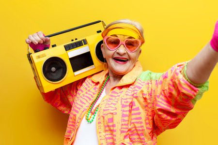 funny crazy granny in hipster clothes listening to music on tape recorder and taking selfie on yellow isolated background, elderly cool woman dancing at party and smilingの写真素材