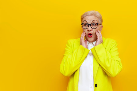 surprised old businesswoman in glasses and formal wear looks away in shock on yellow isolated background, elderly pensioner grandmother in blazer worries in amazementの写真素材