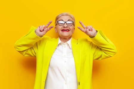crazy old businesswoman in glasses and formal wear showing peace gesture and tongue out on yellow isolated background, elderly pensioner grandmother in blazer showing number four with fingersの写真素材