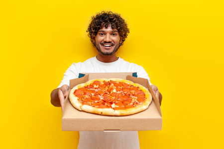 young indian man holding pizza box and showing on yellow isolated background, curly guy student showing and advertising fast foodの写真素材