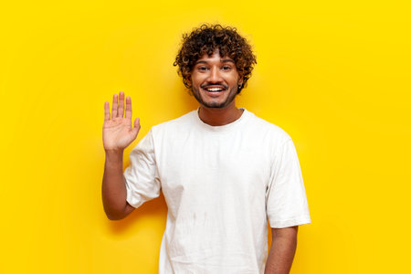 young cheerful indian man in white t-shirt waving and greeting on yellow isolated background, curly guy showing palm gesture welcome and goodbyeの写真素材
