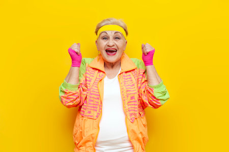 crazy funny old granny in sports colorful clothes wins and celebrates success on a yellow isolated background, elderly woman in youth hipster clothes rejoices in victory and luck and screamsの写真素材