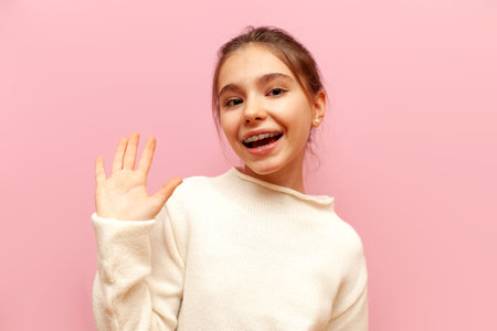 cheerful teenage girl with braces greets and shows palm on pink isolated background, child smiles and shows hello and bye gestureの写真素材