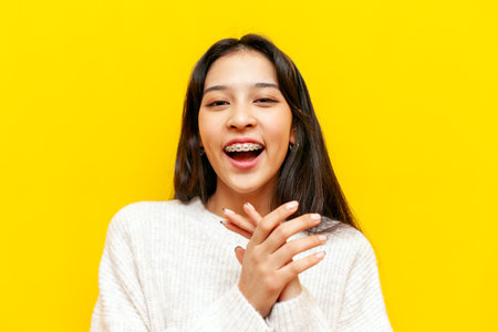 young cheerful asian woman with braces applauding and laughing over yellow isolated background, korean girl clapping and jokingの写真素材