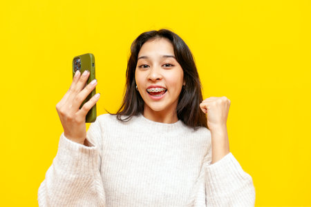 young asian woman with braces using smartphone and winning on yellow isolated background, korean girl winner celebrating success and good luck onlineの写真素材