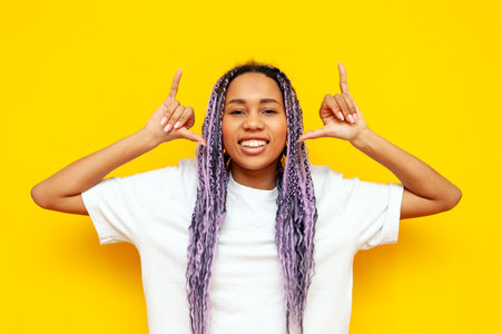 young african american woman with hipster colored dreadlocks smiling and showing youth gesture with hands on yellow isolated background, girl with unique purple hairstyle looking at cameraの写真素材