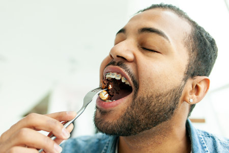 African American man with braces eating chocolate cake with a fork, man holding fork with food and biting with open mouth, close-upの写真素材