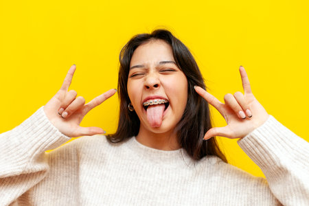 young cheerful asian girl with braces showing rock gesture with hands and tongue on yellow isolated...