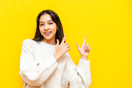 young asian girl with braces points with her hands to the side on a yellow isolated background, korean girl showing and advertising copy spaceの写真素材