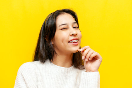 young asian woman with braces winks and smiles on yellow isolated background, korean girl hints and clicks her eyeの写真素材