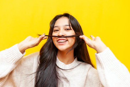 young funny asian woman with braces showing hair mustache on yellow isolated background, korean girl...