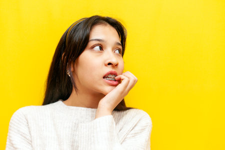 young scared asian girl with braces worrying and biting her paals on yellow isolated background, korean girl confused and puzzled by mistake and problemsの写真素材