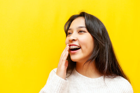 young modest asian girl with braces smiling and taunting on yellow isolated background, korean girl covering her smile and being shyの写真素材