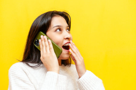 young shocked asian woman with braces talking on phone over yellow isolated background, korean girl...