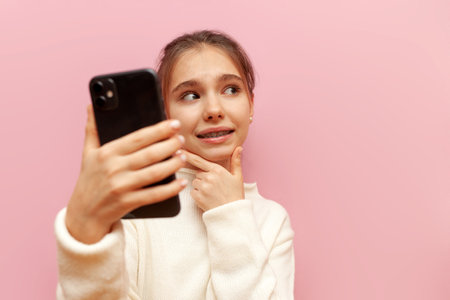 unsure teenage girl with braces uses a smartphone and plans on a pink isolated background, a thoughtful child with a mobile chooses and doubtsの写真素材