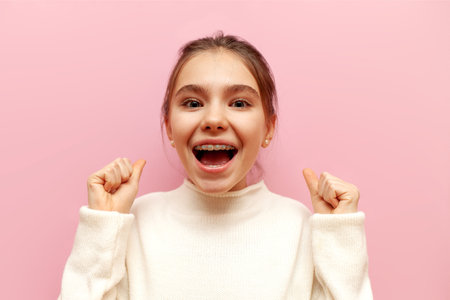 cheerful teenage girl with braces celebrates victory and good luck on pink isolated background, child winner rejoices at success and achievementsの写真素材