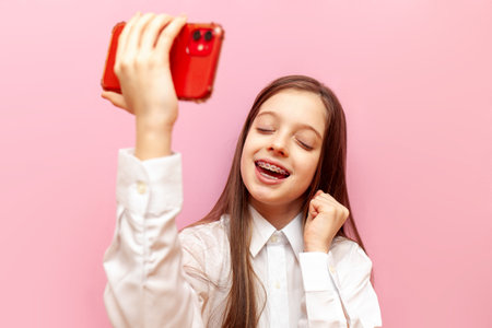 cheerful little teenage girl with braces uses smartphone and rejoices at victory and success on pink isolated background, child winner celebrates good luck online on mobile phoneの写真素材