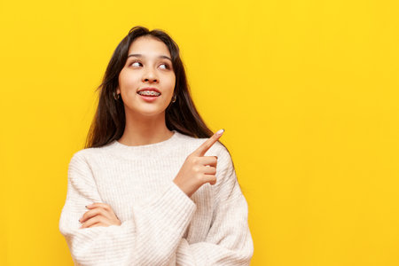 young asian girl with braces points with her hands to the side on a yellow isolated background, korean girl showing and advertising copy spaceの写真素材