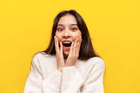 young shocked asian girl with braces wonders on yellow isolated background, amazed korean girl looks at camera with open mouthの写真素材